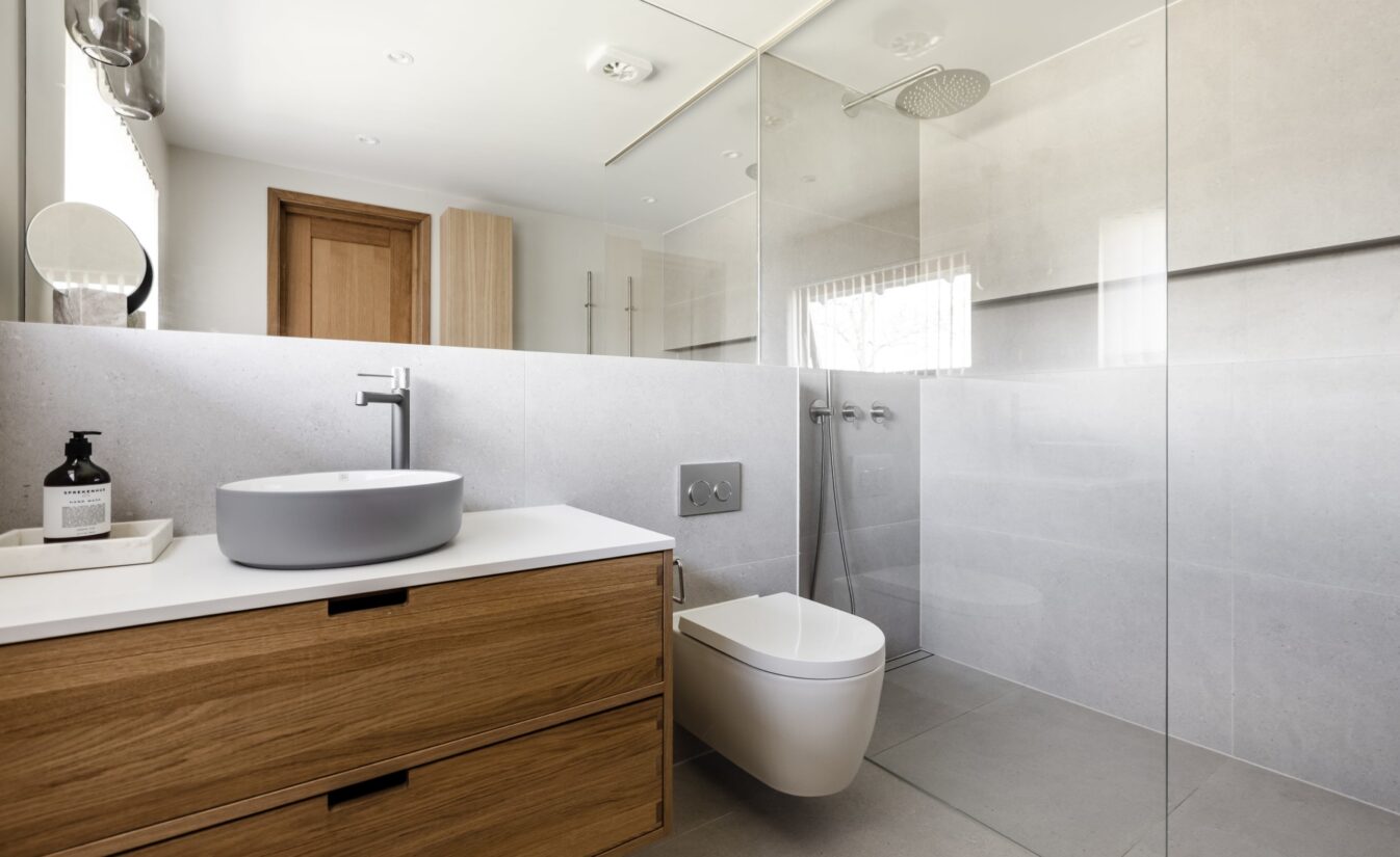 Baderom med store grå fliser, stort dusjrom og møbelment i eik bygget av møbelsnekker. Totalrenovert av Lomundal.