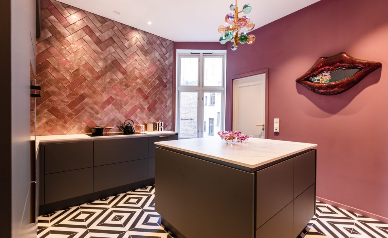 Kjøkken med mørk grå kjøkkeninnredning, geometriske fliser på gulv og rosa vegger. Kjøkkenet er totalrenovert av Lomundal.