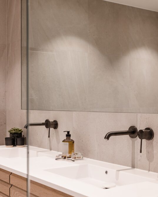 Detaljbilde av baderomsinnredning med dobbel vask og innebygde armaturer i børstet grafitt. Over baderomsmøbelet henger et spesiallaget speil. Utført av Lomundal.