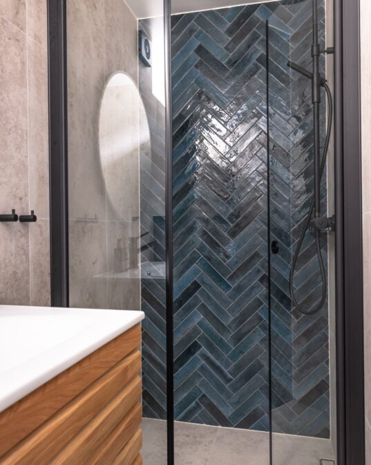 Moderne bad med grå fliser på vegg og gulv, blå blanke fliser i dusj lagt i fiskebensmønster. Armaturer og detaljer i sort finish.
