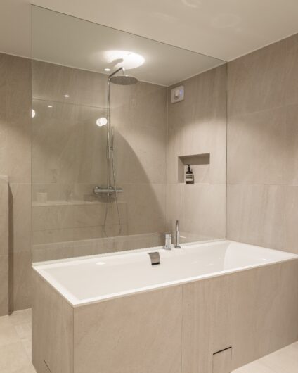 Moderne bad med badekar forran dusjsonen med store beige fliser. Utført av Lomundal.