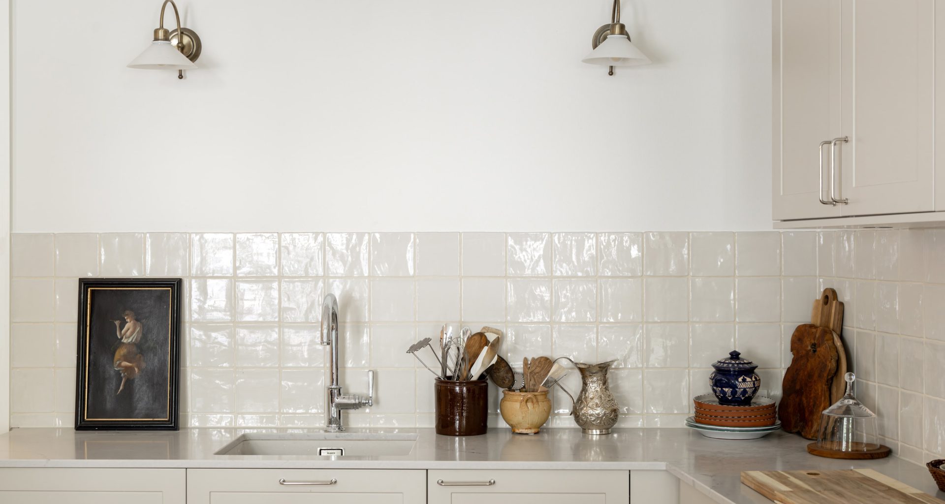 Et lyst og moderne kjøkken i klassisk stil. På veggen er det lagt blanke fliser med ujevne kanter og en bølgete overflate som tilfører personlig preg på kjøkkenet. Utført av Lomundal.