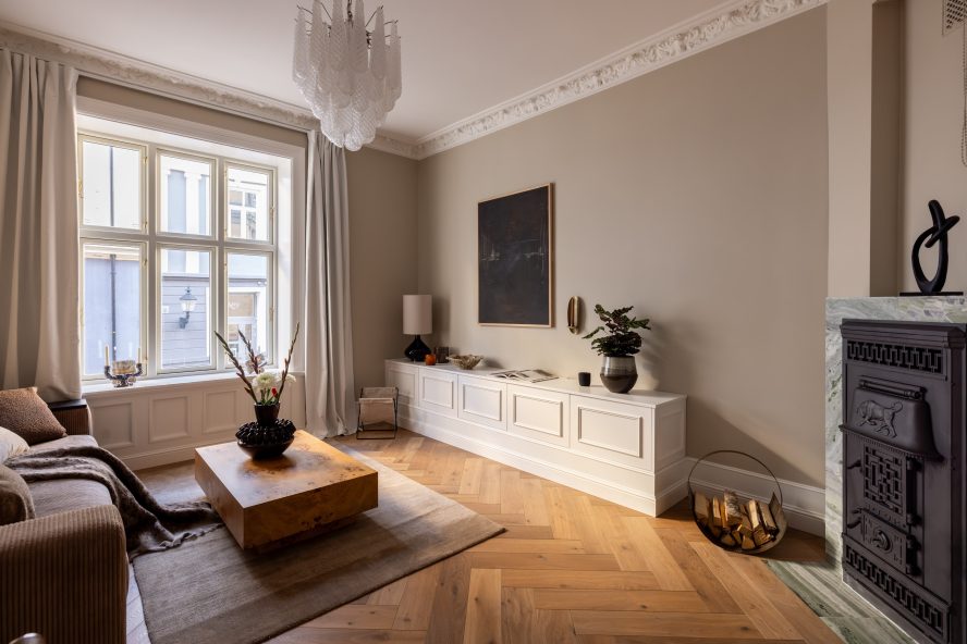 Klassisk stue i beigetoner med elegante stukkaturer, fiskebensparkett og en peis innrammet i blågrønn naturstein. Utført av Lomundal.