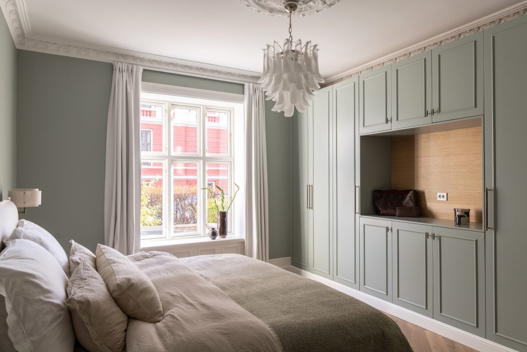 Klassisk soverom i en dus grønnfarge med hvite, elegante stukkaturlister og rosett. Rommet har en behagelig og lys atmosfære, med detaljer i lyst treverk og plassbygd skap. Utført av Lomundal.