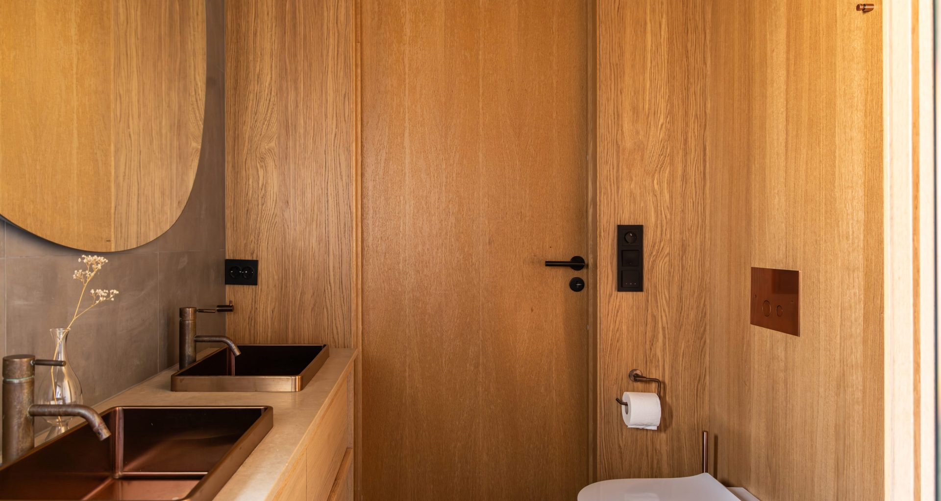 Moderne bad som kombinerer kalde, mørke fliser med varm eikefinér på vegg og baderomsmøbler i mørkt tre. Utført av Lomundal.