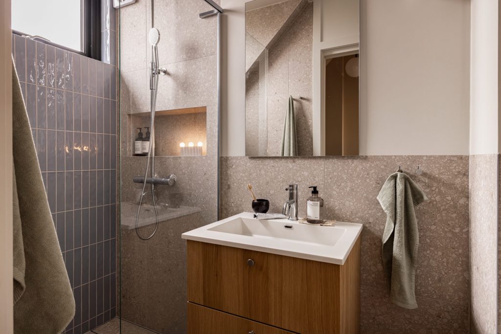 Skandinavisk bad med blå, blanke fliser i dusjsonen, terrazzoflis på gulv og vegger og baderomsinnredning i lyst treverk med armaturer i krom. Badet er totalrenovert av Lomundal.