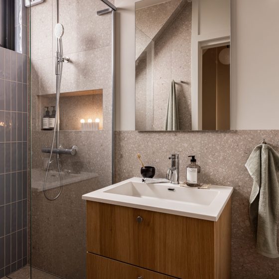 Skandinavisk bad med blå, blanke fliser i dusjsonen, terrazzoflis på gulv og vegger og baderomsinnredning i lyst treverk med armaturer i krom. Badet er totalrenovert av Lomundal.