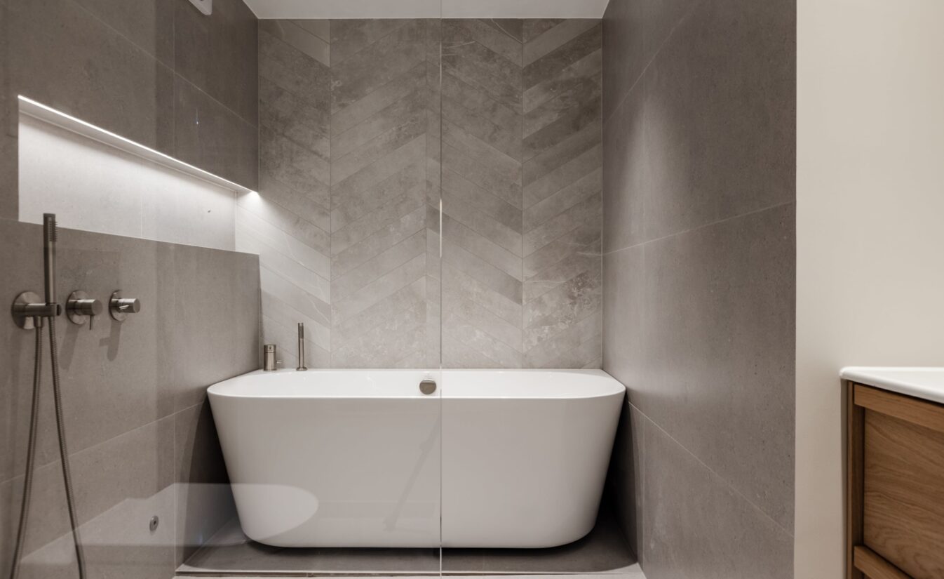 Detaljbilde fra bad av dusjrom og badekar med store grå fliser på gulv og vegg, med egne chevron fliser bak badekaret. Utført av Lomundal.
