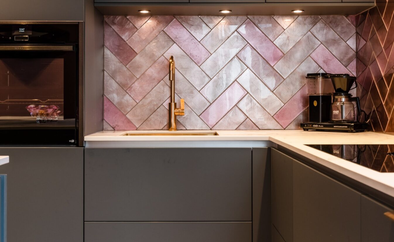 Kjøkken med grå kjøkkeninnredning, geometriske fliser på gulv, chevronfliser i rosa på vegg og vask og armatur i messing. Utført av Lomundal.
