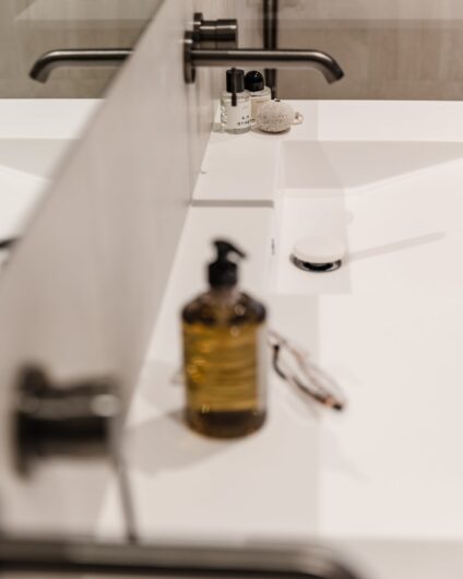 Detaljbilde av dobbel vask med innebygde armaturer i børstet grafitt. På vasken står parfymeflasker, håndsåpe og noen briller.