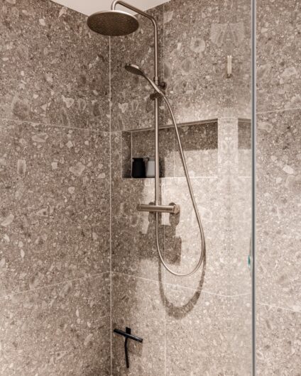 Detaljbilde av dusjrom med armaturer i børstet stål. Flisene på gulv og vegg er grå med mye spill i. Designsluk fra Purus og plassbygd hyllenisje til baderomsartikler. Utført av Lomundal.