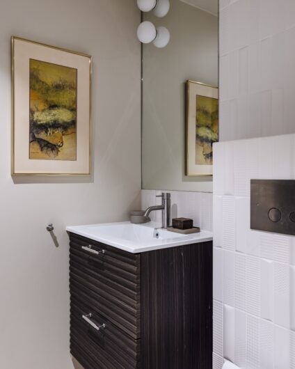 Toalettrom med flislagt vegg over toalett og ellers malte flater. Baderomsinnredning i valnøtt med lamper på speilet over.