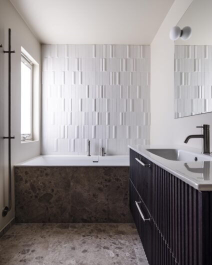 Moderne baderom med fliser på gulv og rundt badekar i beige stein. Over badekaret er det lagt 3D dimensjonale fliser. Utført av Lomundal.