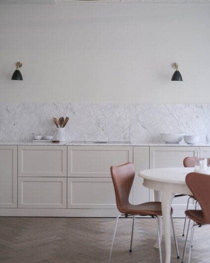 Beige og klassisk kjøkken fra Drømmekjøkkenet med beige fronter og marmor benkeplate. Sort og messing veggehengte lamper over kjøkken. Lyst spisebord med skinnstoler fra Fritz Hansen.