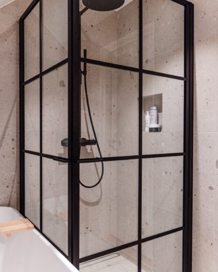 Dusj på bad med beige fliser. Dusjdørene har sorte rammer som står i stil med dusjarmaturet i matt sort. Utført av Lomundal.