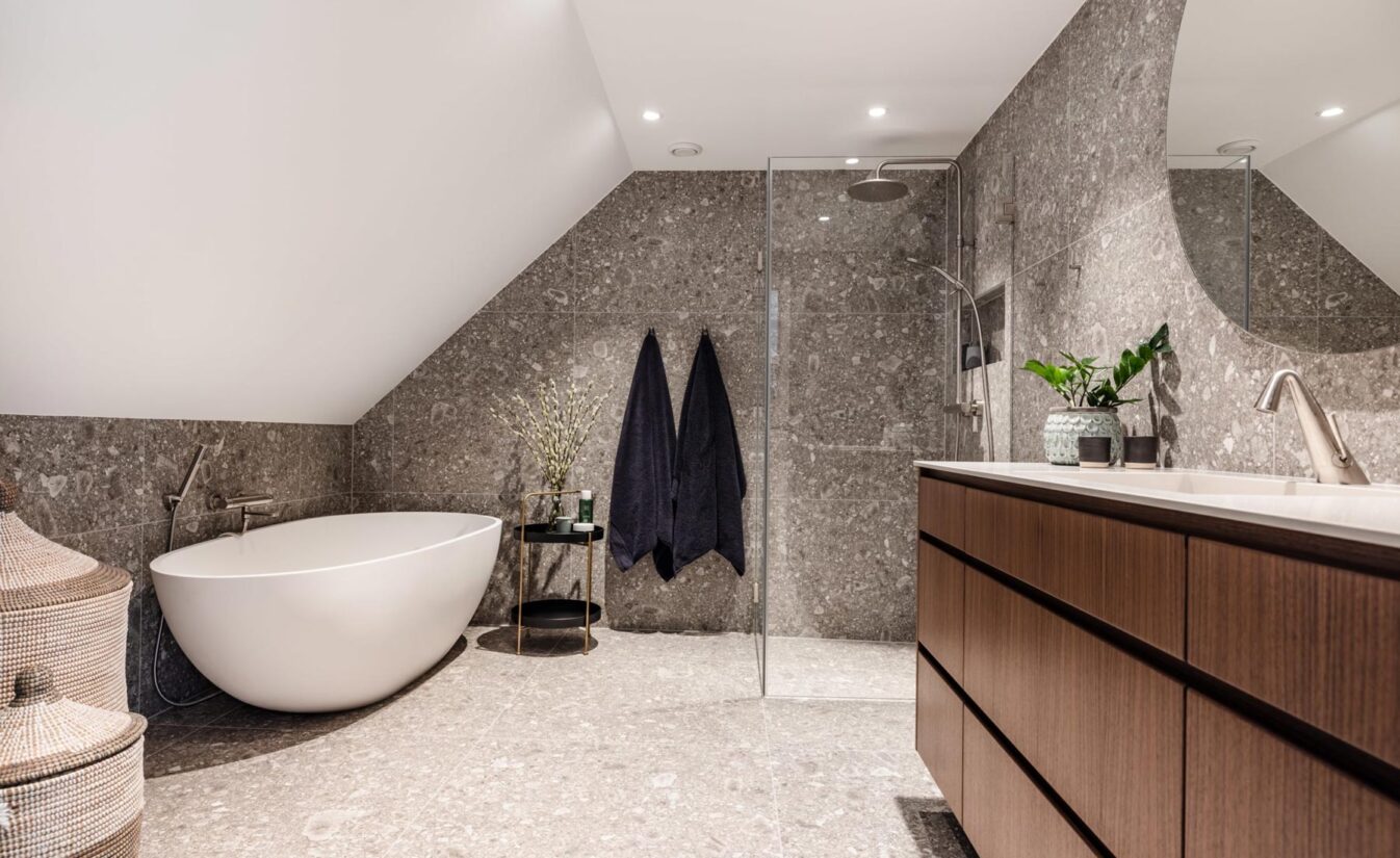 Baderom med store grå fliser, frittstående badekar og baderomsinnredning i mørk treverk. Utført av Lomundal.