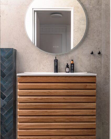 Moderne bad med grå teksturerte fliser på gulv og vegg, baderomsinnredning i eik og rundt speil fra Linn bad. Utført av Lomundal.