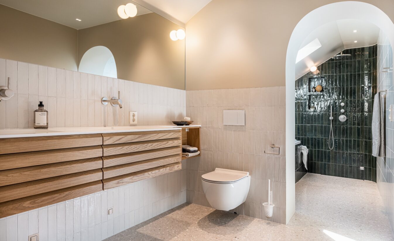 Bad med blanke loddrette fliser på vegg, terrazzoflis på gulv og baderomsinnredning i eik. Utført av Lomundal.